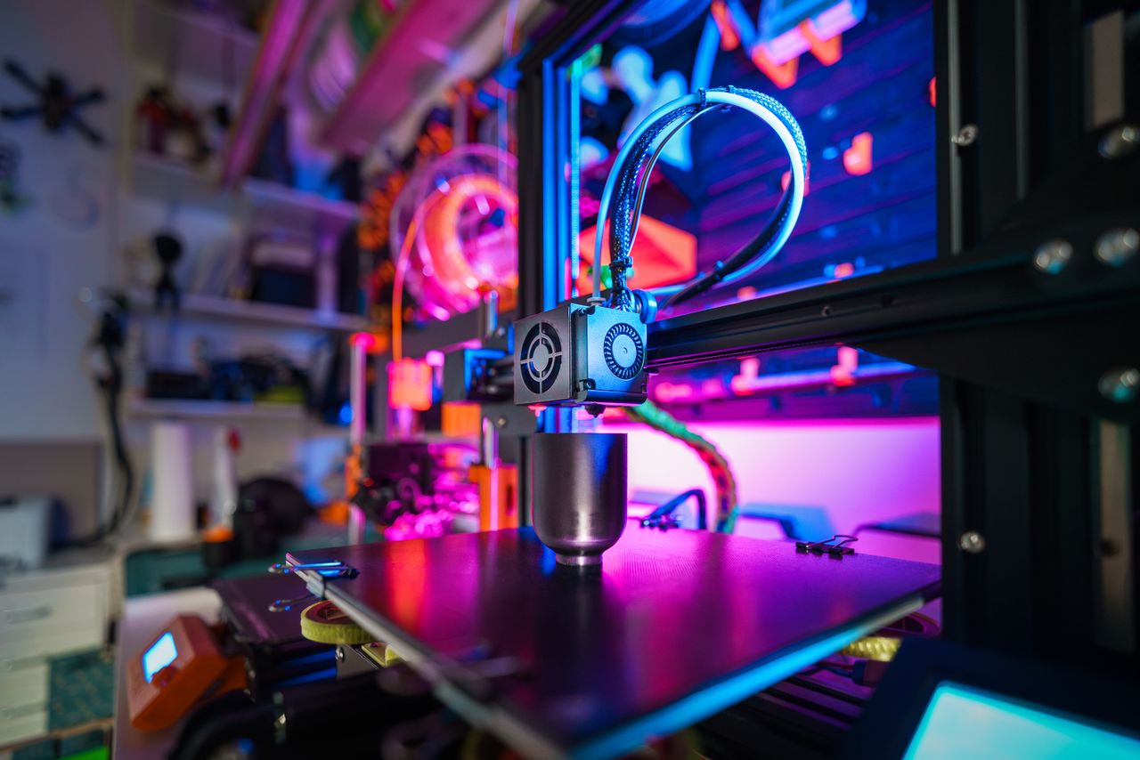 about-img-02 A close-up of a modern 3D printer in action, illuminated by vibrant, colorful lighting indoors.